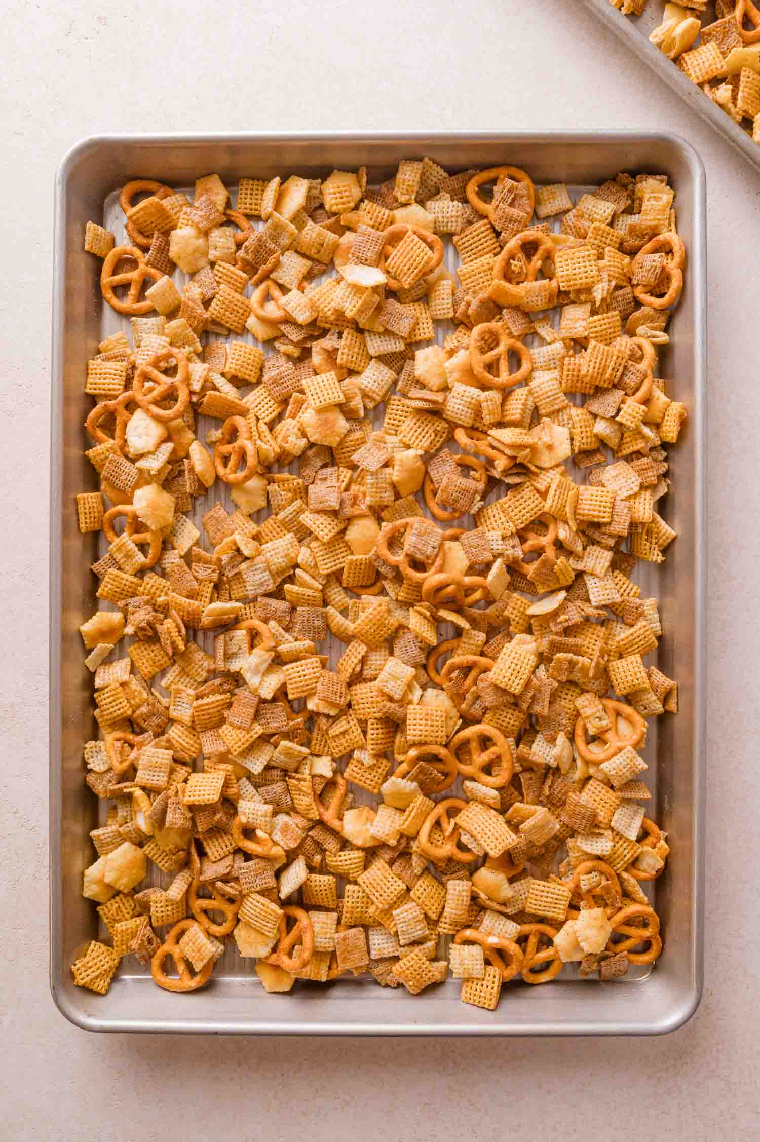 A metal baking sheet filled with a snack mix of square cereal pieces and pretzels.