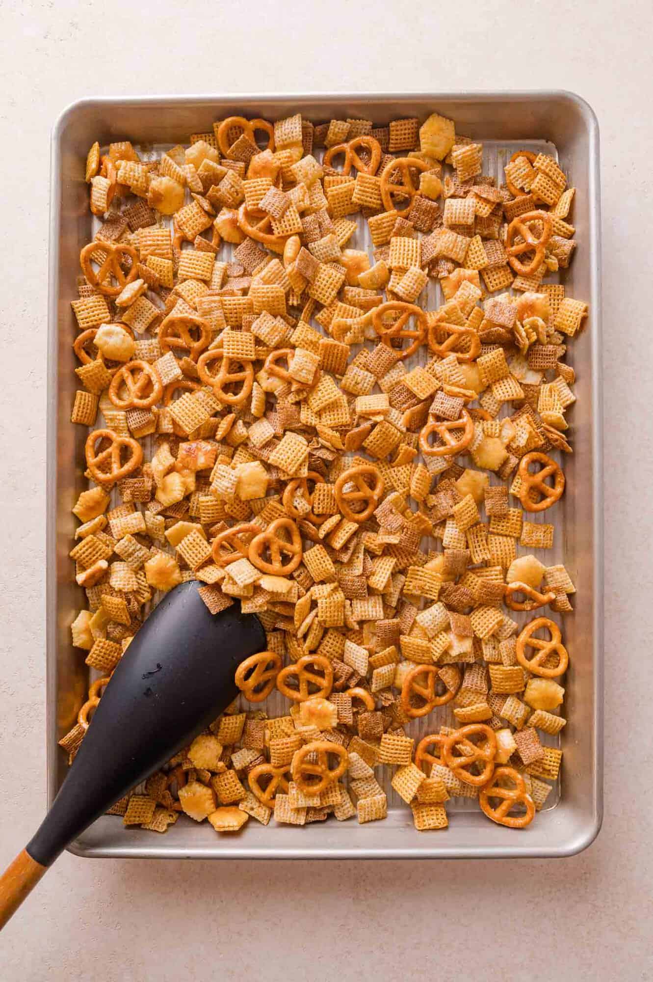 A baking sheet filled with a hot honey chex made with a mix of cereal squares and pretzels, with a black spatula scooping a portion.