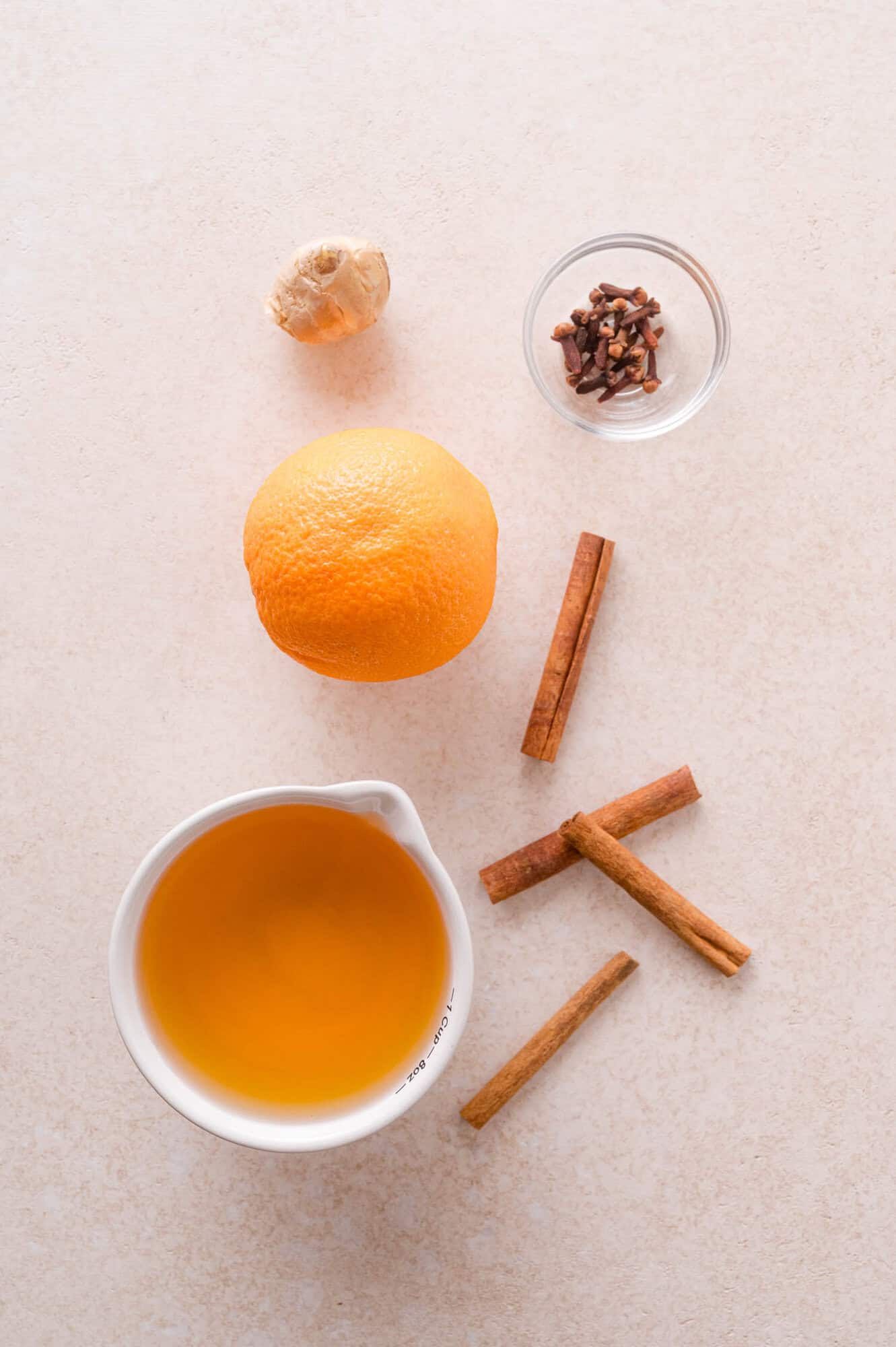 An orange, a piece of ginger, a small bowl of cloves, three cinnamon sticks, and a measuring cup filled with apple cider.