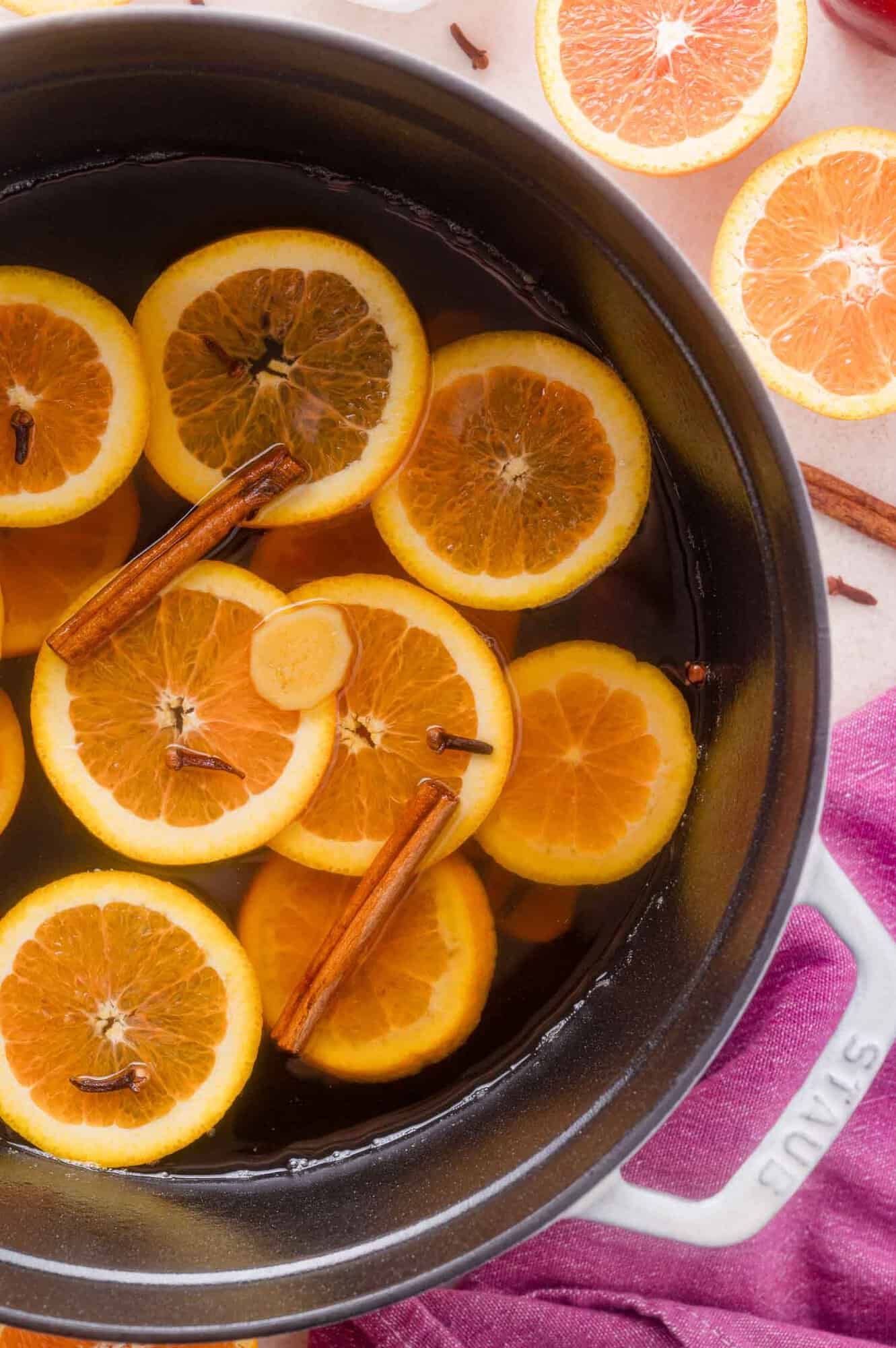 Pot of mulled apple cider containing orange slices, cinnamon sticks, and cloves, with additional orange slices nearby.