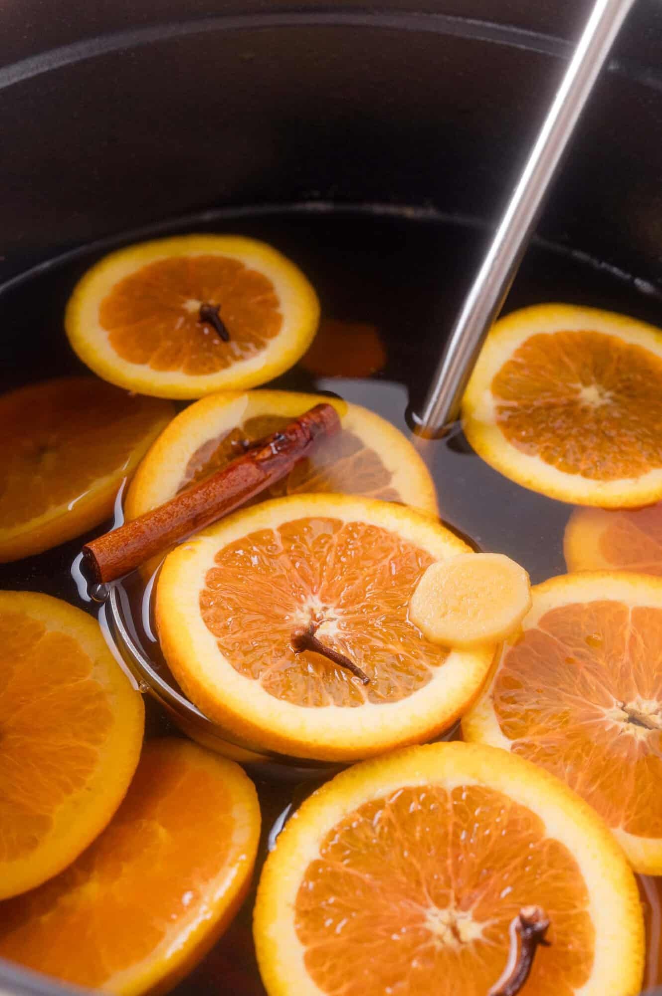 A pot of apple cider filled with orange slices, a cinnamon stick, cloves, and a piece of ginger, with a metal ladle.