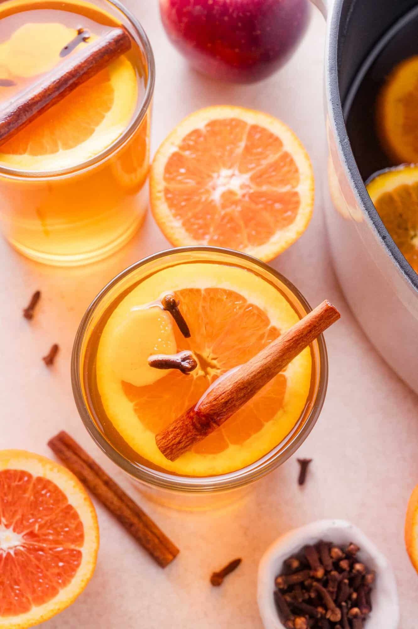 Two glasses of mulled cider garnished with orange slices, cinnamon sticks, and cloves, next to a pot of cider and fresh fruit.