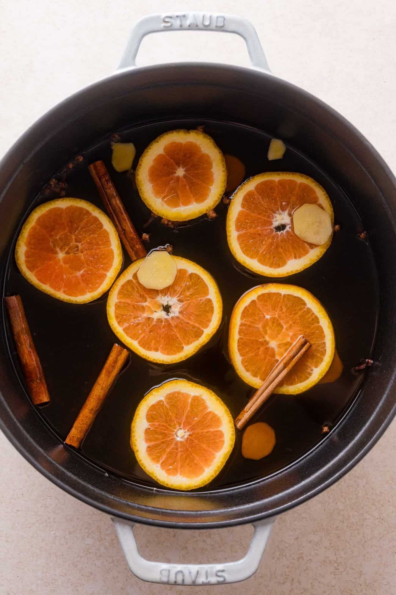 A Staub pot filled with apple cider containing orange slices, cinnamon sticks, ginger pieces, and cloves.